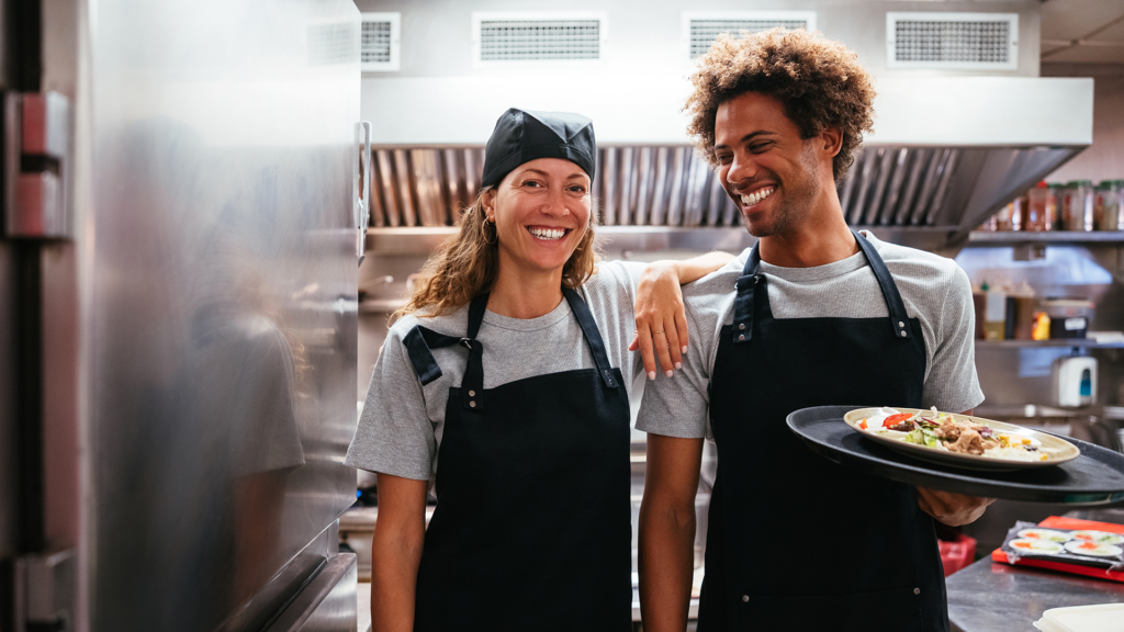 Image shows two colleagues standing in the kitchen of a restaurant. There are stainless steel appliances around them. They are both wearing a uniform of a light gray t-shirts and black aprons. One person is female presenting with golden skin and long, light brown wavy hair. She is wearing a black kitchen prep hat over her hair. She is smiling into the camera and has her hand on her colleague's shoulder. The other is male presenting with light brown skin and natural, curly hair. He is holding a tray with a plate of food on it and smiling at her.