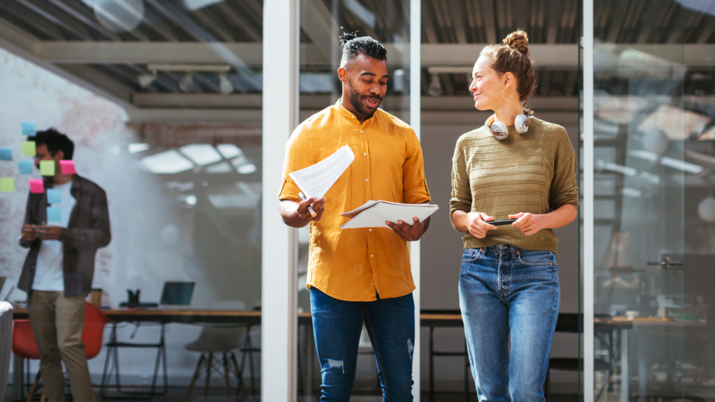 Two colleagues standing and comparing notes after a meeting. They are both dressed very casually. The male presenting person is Black and is wearing an orange button down with dark blue jeans. His hair is shaved on the sides and longer on top and he has some facial hair. He is holding a notebook and some loos leaf papers in his hands. The female presenting person is white. Her light brown hair is pulled up into a bun and she has a pair of silver headphones around her neck. She is wearing a green sweater with lighter blue jeans. They are smiling at each other and appear to be having a pleasant conversation.