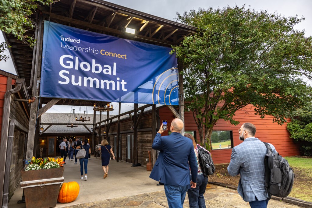 Several attendees enter the modern barn-like venue under a blue banner reading Indeed Leadership Connect Global Summit, the surroundings decorated by potted plant arrangements and pumpkins.