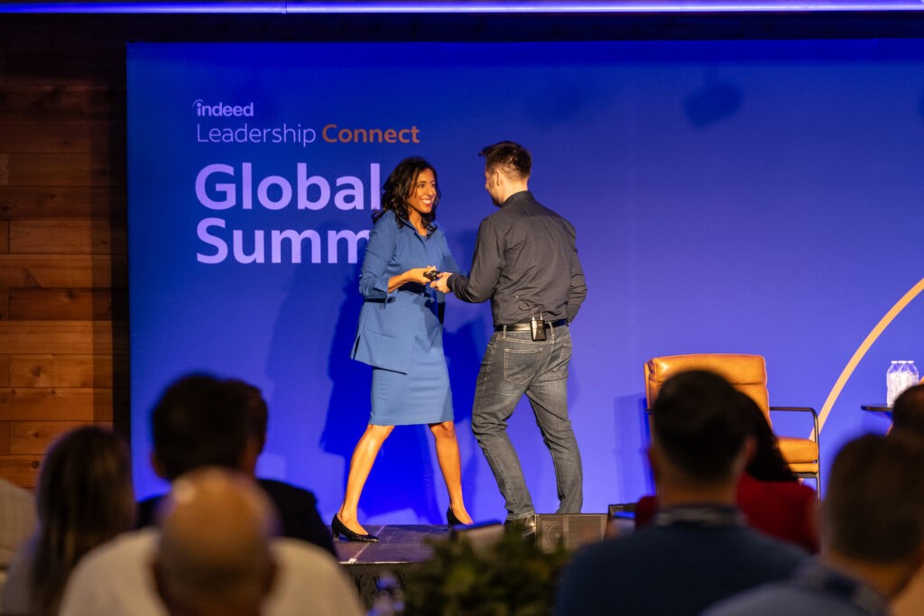 On a stage in front of the audience, Erica Dhawan, in a baby blue blazer and matching dress, dances and laughs with Indeed Senior Director of Corporate Marketing Daniel Wahlen, who wears gray jeans and a black collared shirt.
