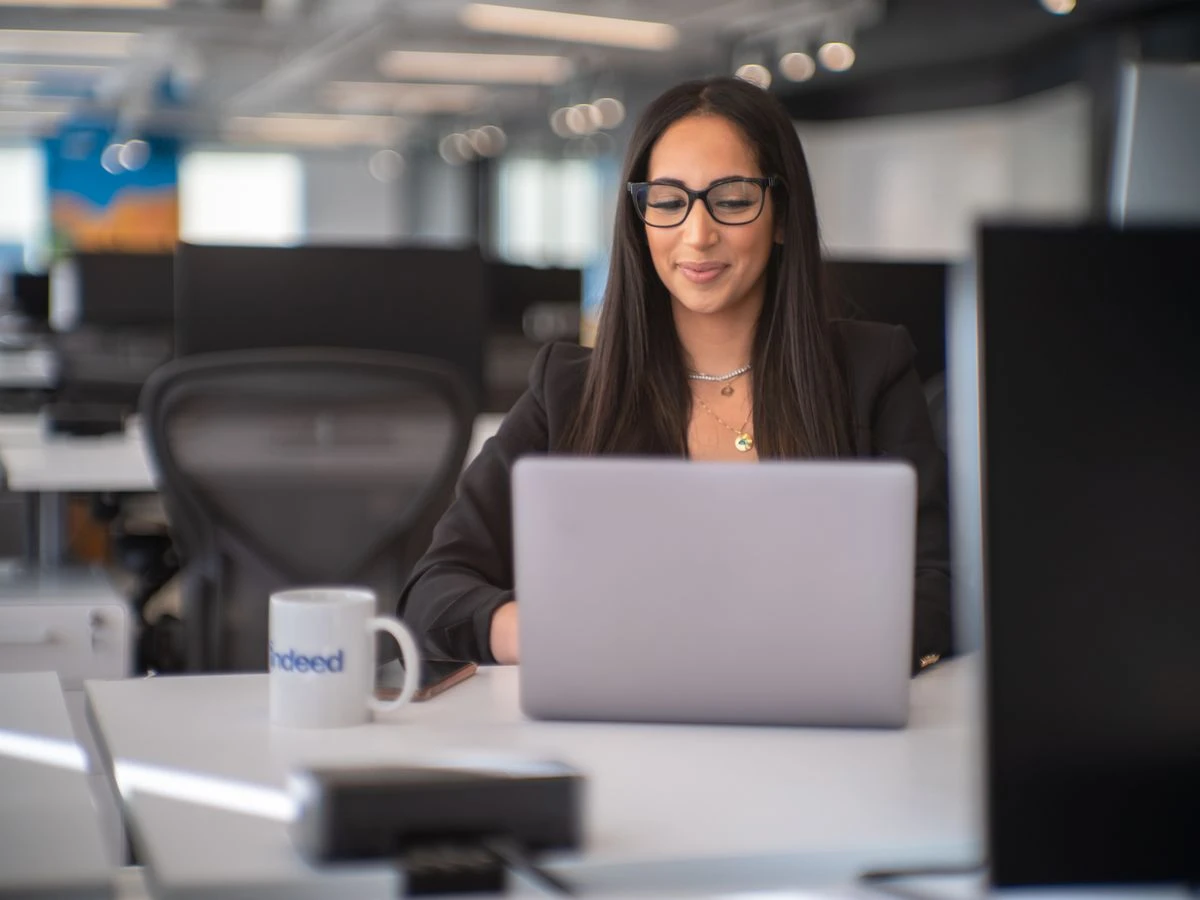 Person wearing glasses working on a laptop in an office setting with an 'Indeed' branded mug on the desk.