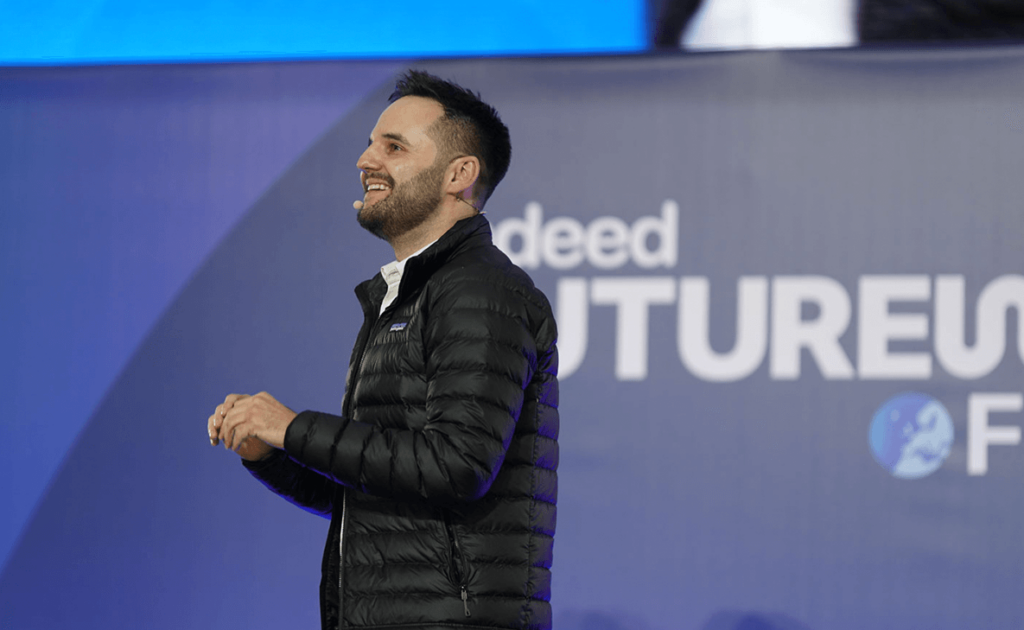 man presenting on stage wearing a black jacket and a head set in front of a blue Indeed Futureworks banner