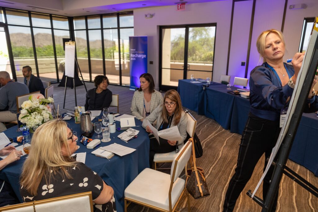 A group of Recharge attendees take part in a discussion and brainstorming exercise. One group sits at a round table in the conference room sharing notes and talking while one blonde woman in dress pants and a blue blouse stands and writes notes on an easel to share later with the larger group.
