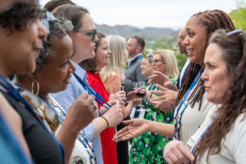 A group of Recharge attendees stand in a line facing each other during a conversational exercise at Recharge. They are outdoors in a garden with a mountain range visible in the background, smiling and gesturing excitedly as they converse.