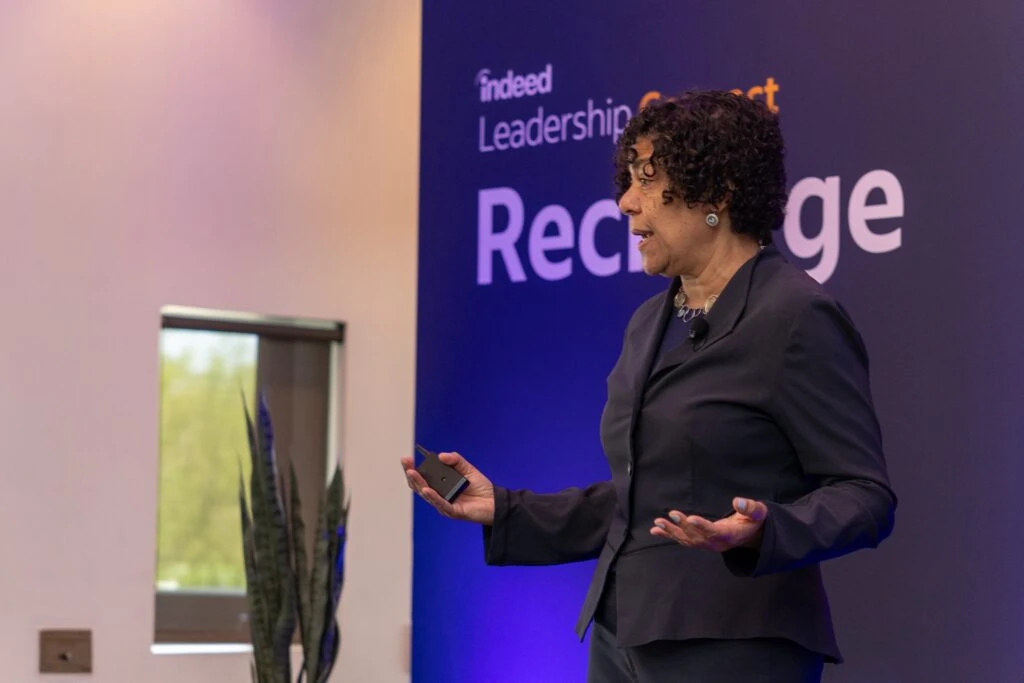 Dr. Heidi Brooks speaks on the Leadership Connect Stage, a banner reading Indeed Leadership Connect Recharge behind her. She has dark curly hair cropped close to her head, a dark grey blazer and bauble earrings and a matching necklace. She is gesturing animatedly with her hands while she speaks.