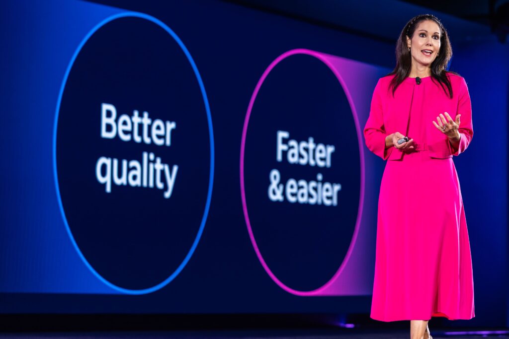 Indeed Chief Revenue Officer Maggie Hulce speaks in front a presentation screen wearing a hot pink dress and matching jacket, with her long brunette hair held back with a braidd headband. The screen behind her reads, "Better quality" and "faster and easier."