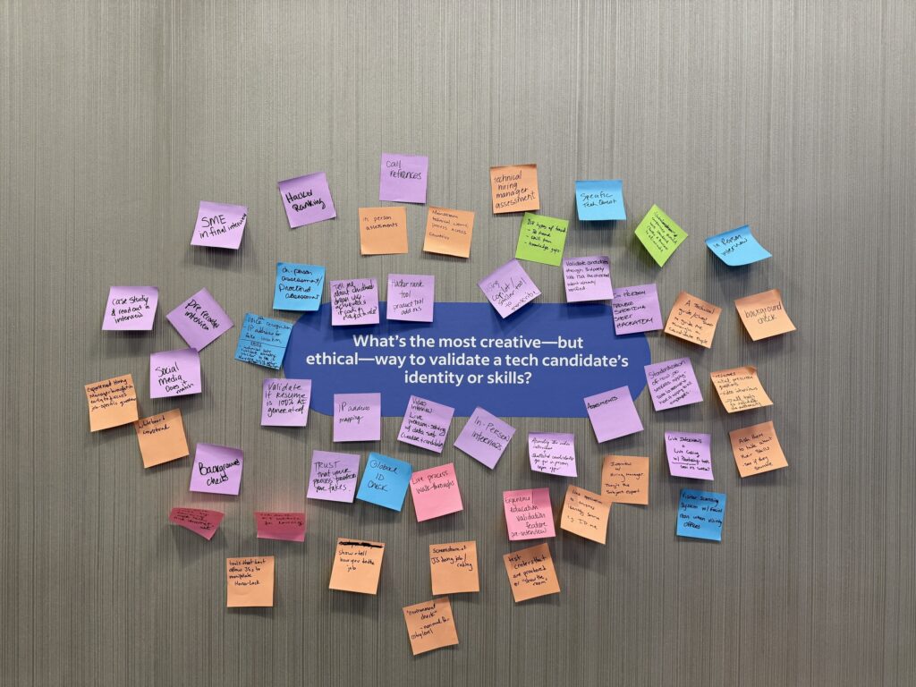 On a wall are colorful sticky notes surrounding a purple paper with the question: What’s the most creative but ethical way to validate a tech candidate’s identity or skills? The numerous, handwritten sticky notes from employers include answers like technical hiring manager assessments, background checks, and having the candidate bring a case study demonstrating their experience to an interview.