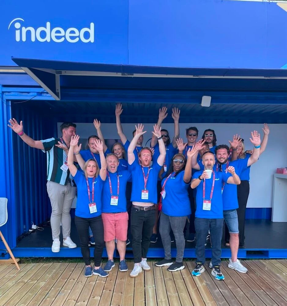 A cheerful group of 15 Indeedians posing in front of a blue Indeed branded booth at an event.