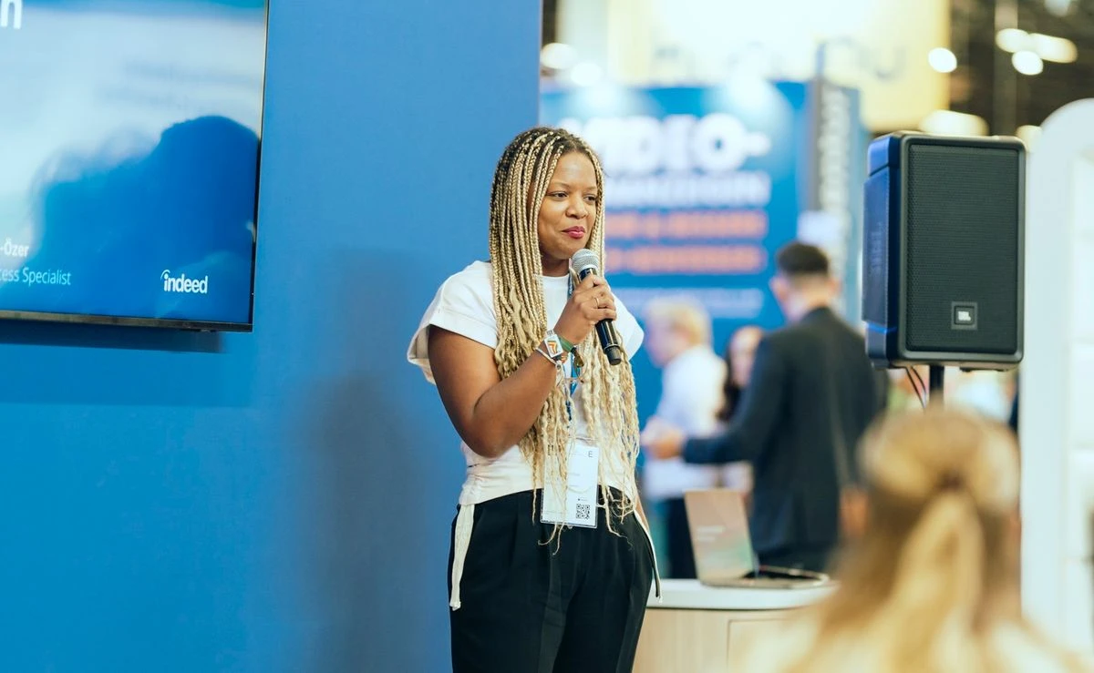 Serah speaking into a microphone at a trade show, standing in front of a blue Indeed branded booth