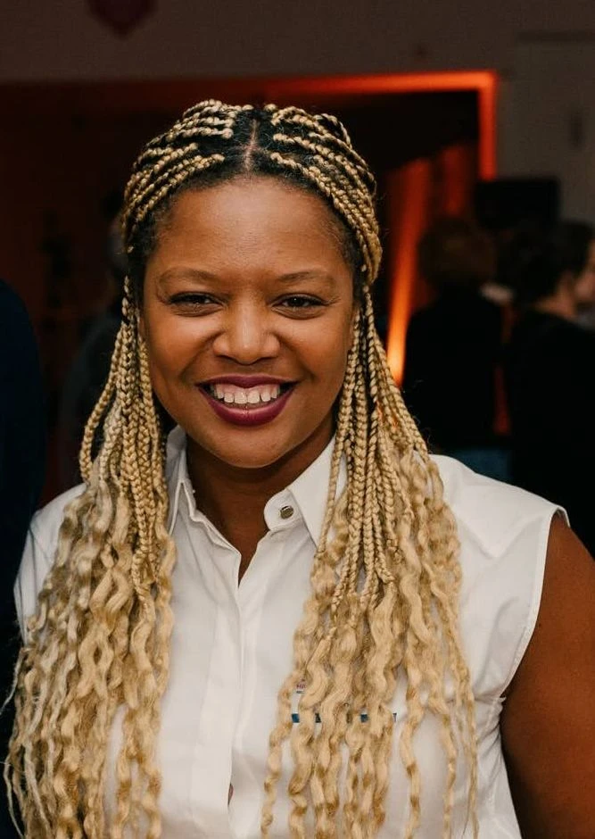 Serah smiling with long braided hair, wearing a white sleeveless shirt and beige pants. They are in a social gathering environment at the Indeed HR slam event