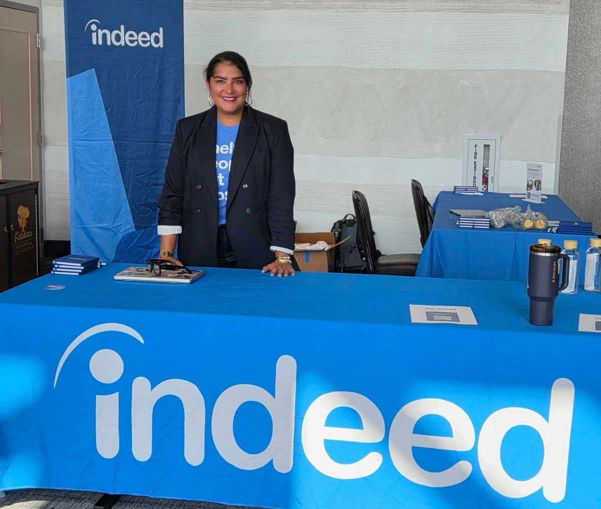 A person stands behind an Indeed-branded table at an event setup, smiling in front of a blue Indeed banner.