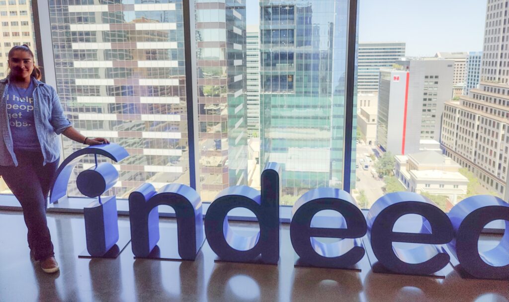 An individual stands next to a large Indeed sign in front of floor-to-ceiling windows overlooking downtown Austin.