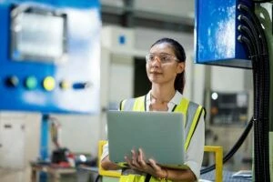 Measure the Productivity, Effectiveness, or Efficiency of Machinery. Female Engineer checking an automated mechanical press machine while standing and input data on a laptop in a production line of the manufacturing industry.