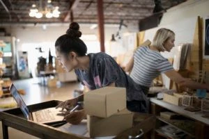 Female small business owner packaging merchandise at laptop in workshop