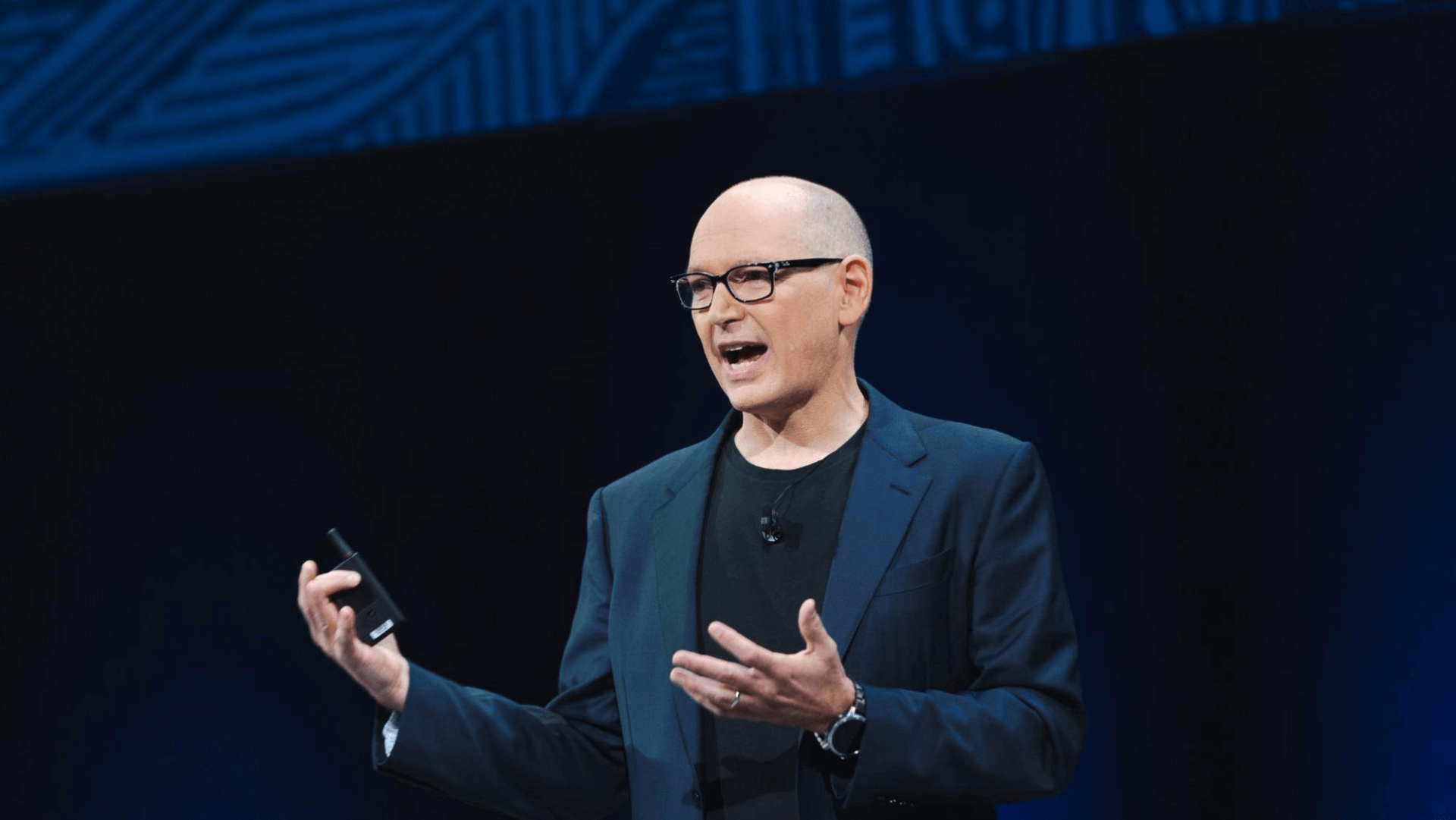 White man presenting during a tech presentation. He is wearing a dark blue blazer with a black tshirt underneath. He has black, thick rimmed glasses on and is holding a clicker in his right hand.