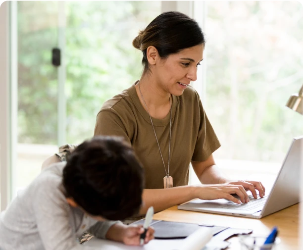 Woman with dark hair sitting at a desk working on a computer next to her child.