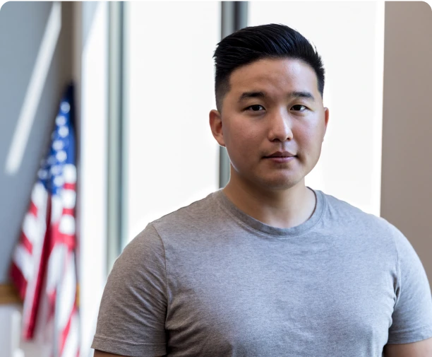 Asian man in gray t-shirt standing in front of a window and American flag.