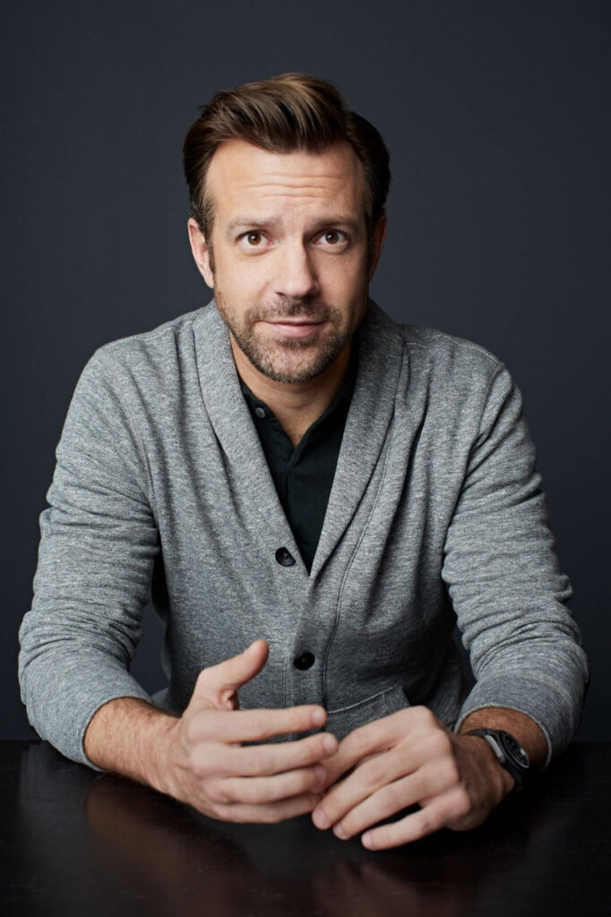 Image of Jason Sudeikis, wearing a gray cardigan, sitting and looking into the camera. 