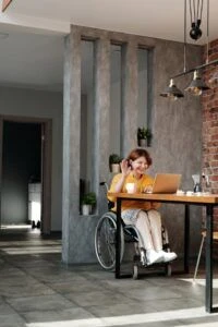 Woman in wheelchair sits at table in front of laptop waving.