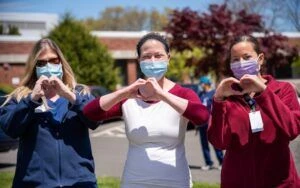 Three healthcare professionals standing outside with masks and forming heart signs with their hands.