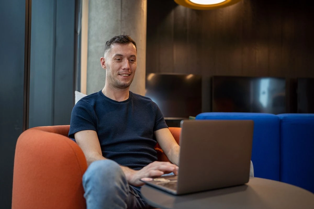 A man with short dark hair and a dark blue t-shirt sits in an orange armchair, smiling slightly as he looks at a laptop on a small table in front of him.
