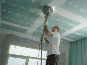 A man sanding drywall on a ceiling in an unfinished room.
