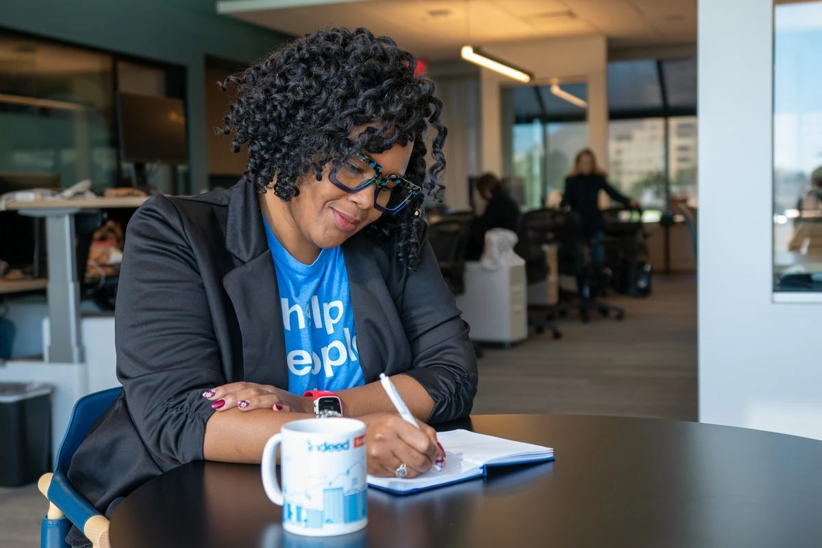 A woman with curly hair and blue glasses is seated at a black round table, writing in an open notebook with a pen.
