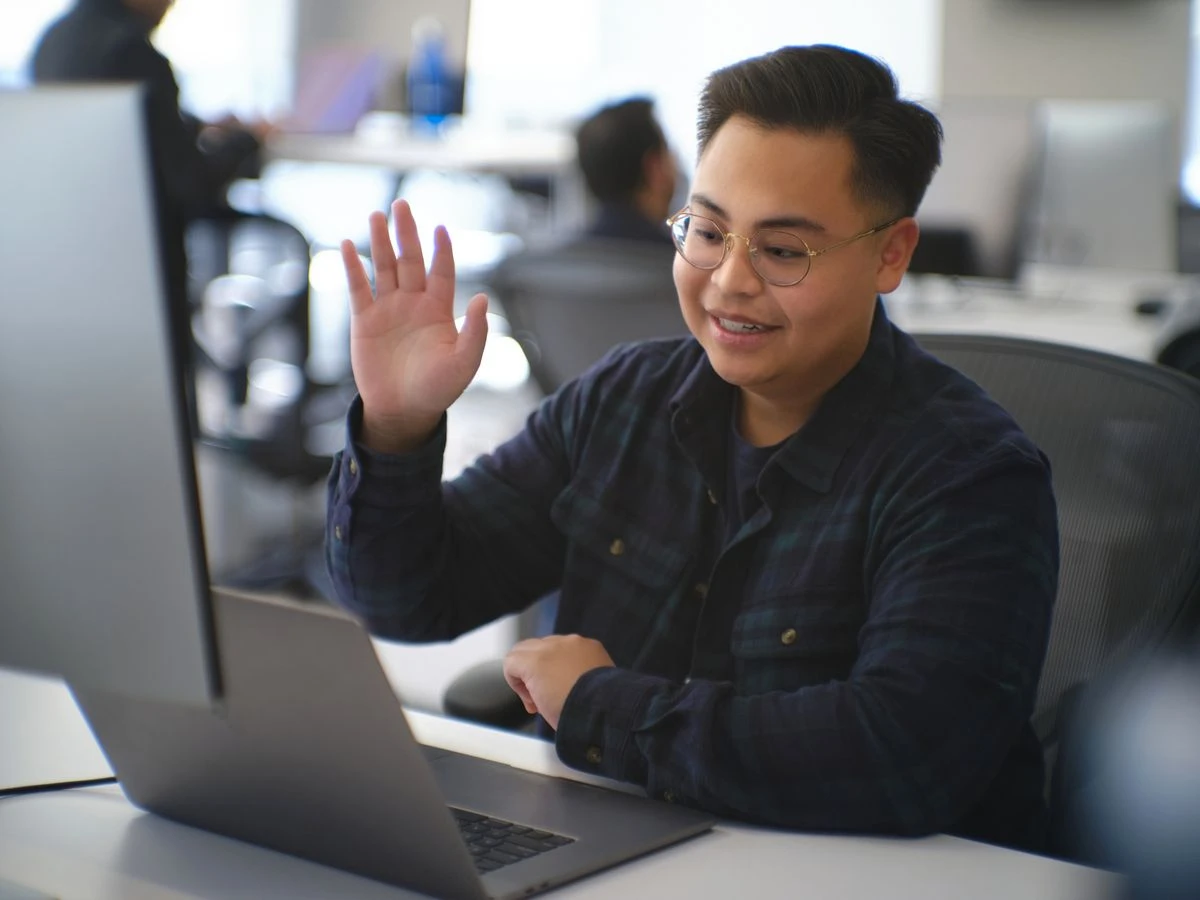 A person with short, dark hair and round glasses sits at a desk in an office, smiling as they wave at a laptop screen. They are wearing a dark blue plaid shirt over a t-shirt.
