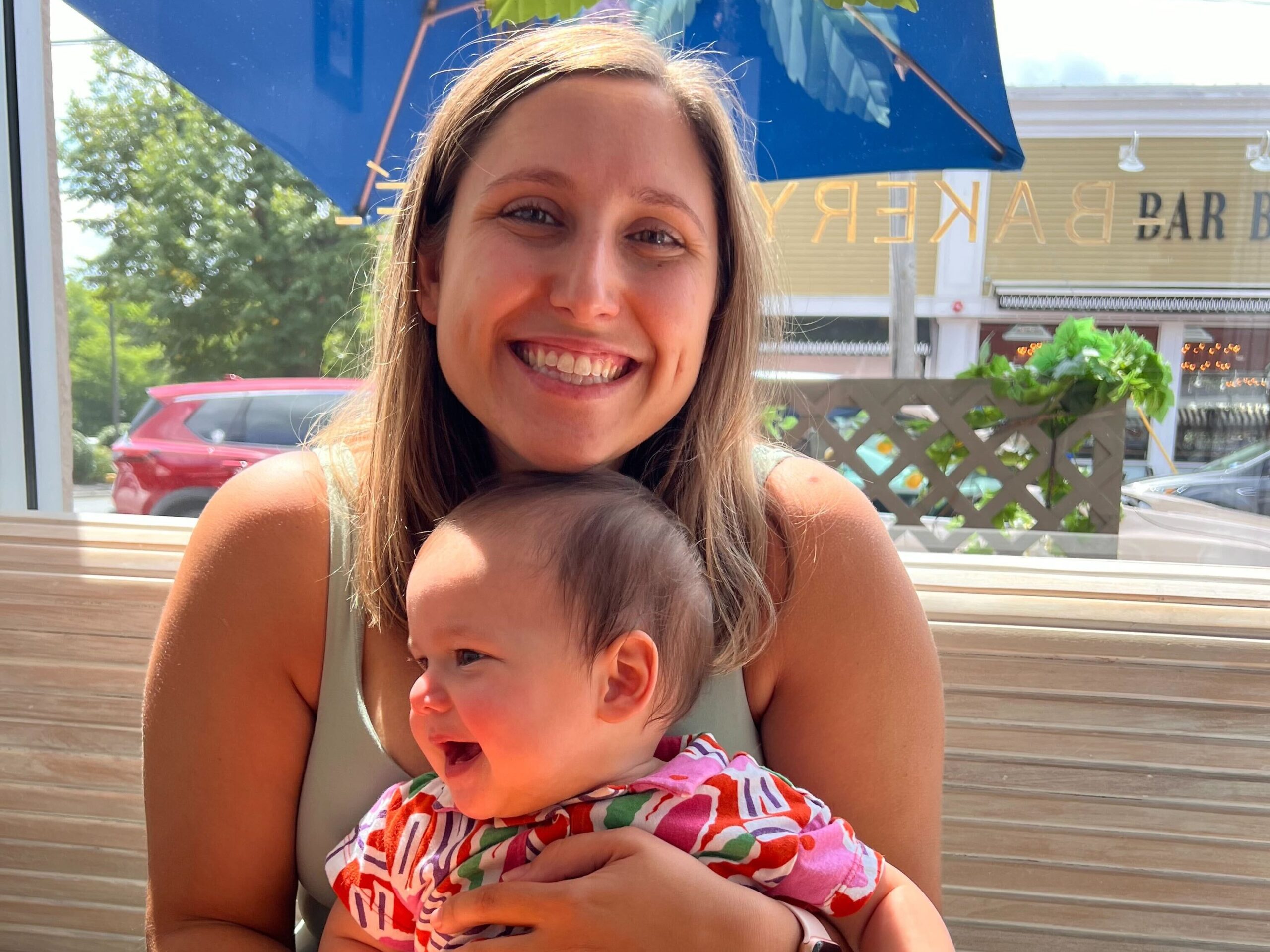 An adult smiling at the camera while holding a joyful baby at an outdoor restaurant under a blue umbrella