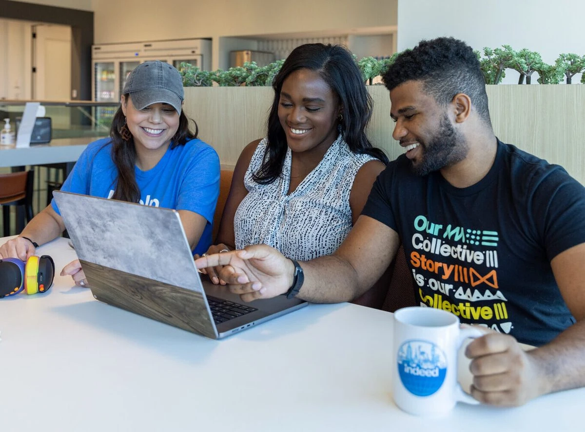 Three people collaborate over a laptop at a table in a modern office with stylish lighting fixtures.