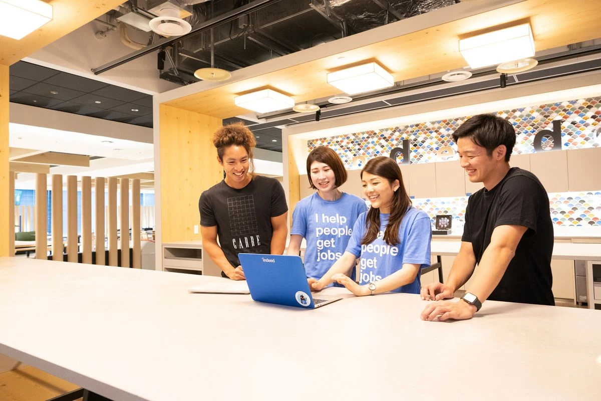 Four people standing at a table in an office setting, using a laptop with a visible Indeed logo on its screen, and engaging in a collaborative discussion
