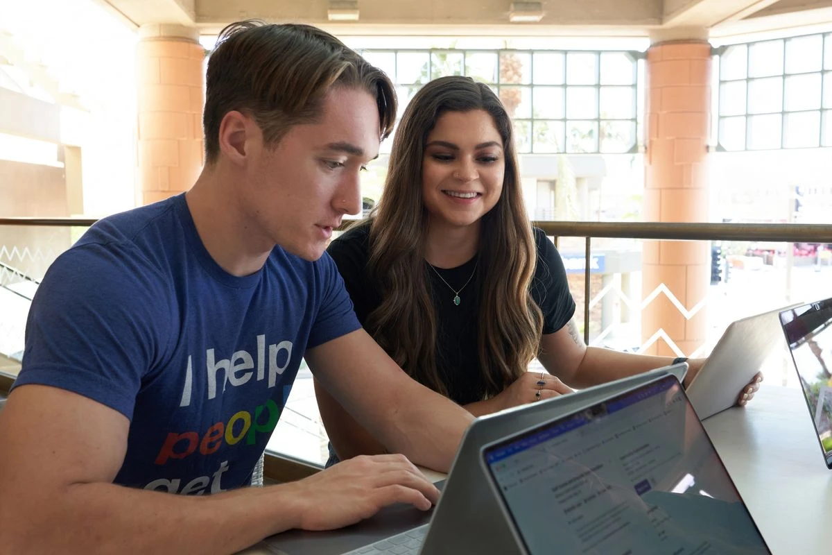 Two people sitting at a table, looking at a laptop screen together in a brightly lit terrace setting. One person is wearing a blue t-shirt with the text I help people get jobs visible