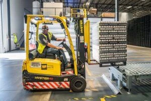 A man driving a forklift in a warehouse, carrying a palette of products.