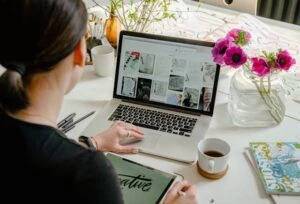 A woman working at her desk on a laptop with a tablet, surrounded by colorful books and flowers.
