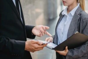 A businesswoman holding an open folder while talking to a businessman holding a phone.