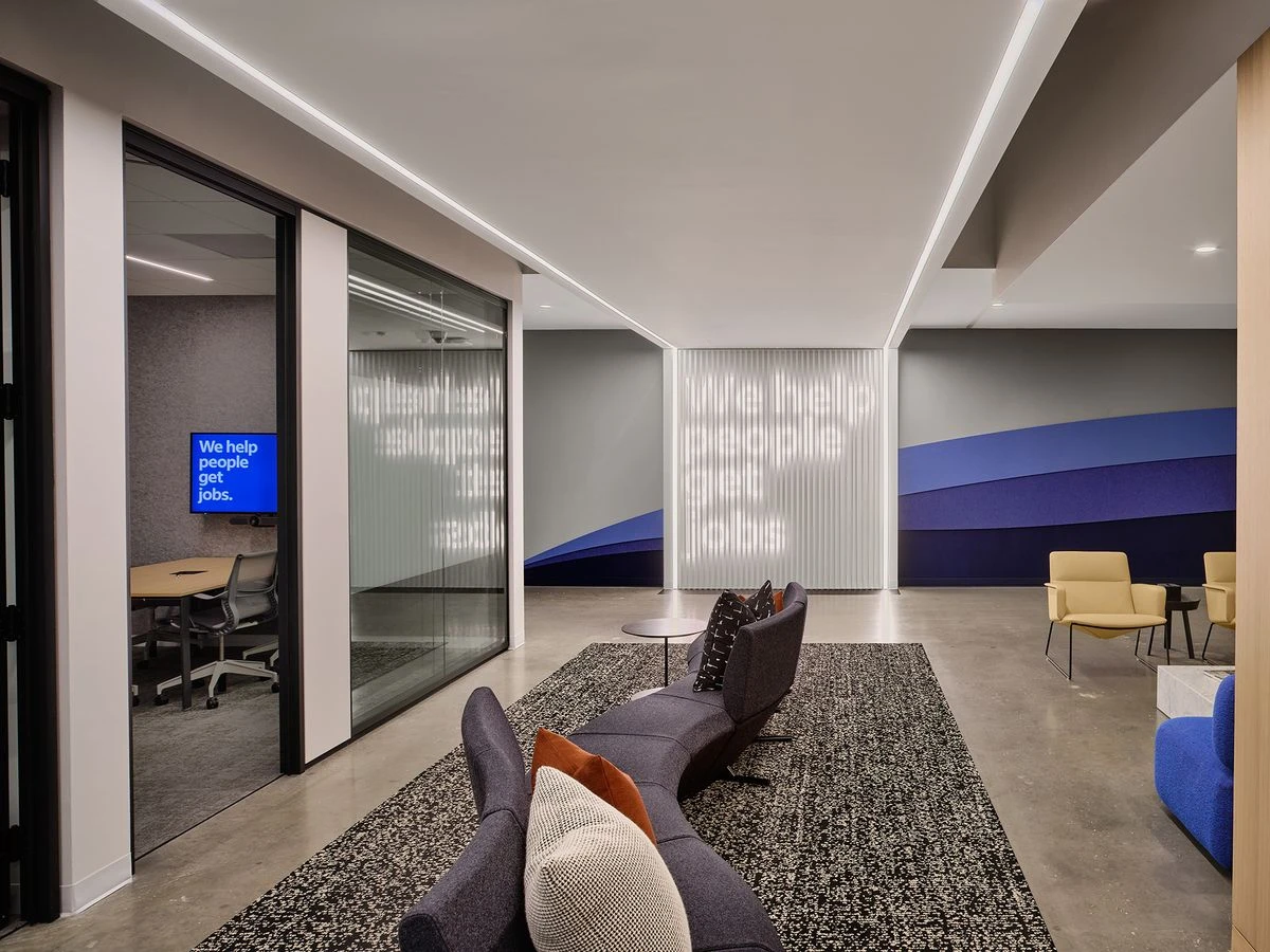 Modern office lobby with sofas and chairs, featuring a blue and gray color scheme. Includes a view of a conference room through glass walls with the text We help people get jobs on the display inside.