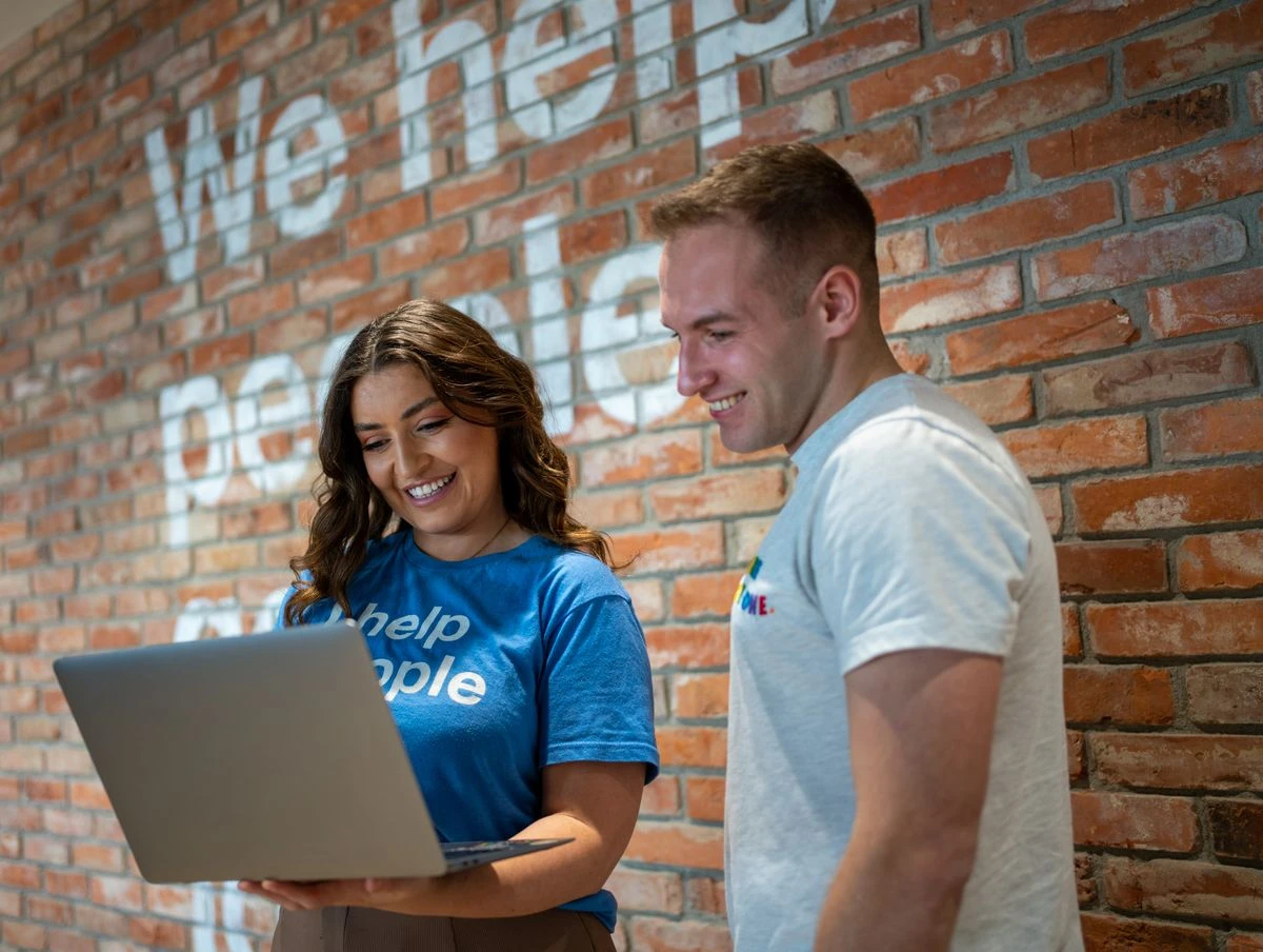Two people smiling and looking at a laptop in front of a brick wall with We help people get jobs in the background