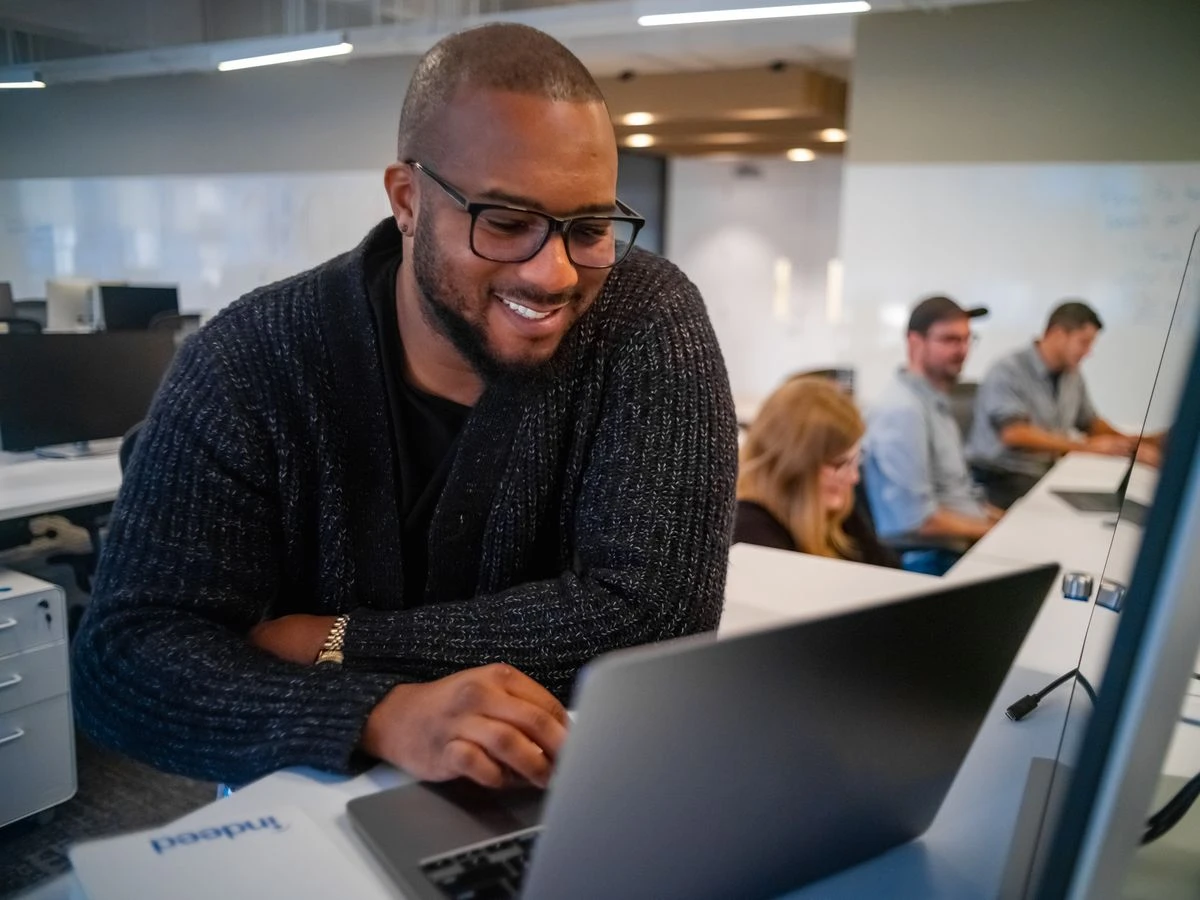 A person smiling while using a laptop in a busy office environment. Other colleagues are focused on their work in the background