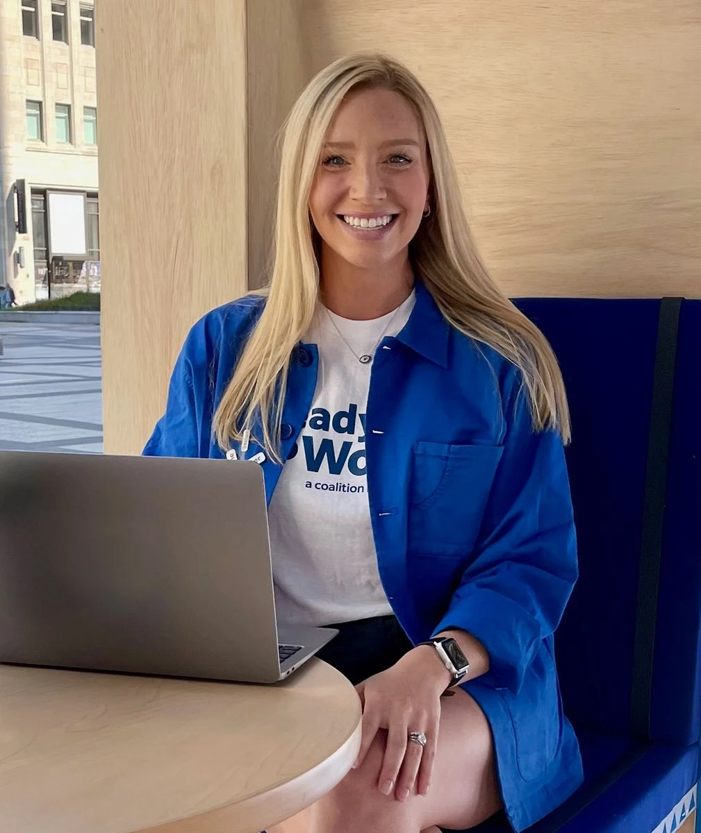 Person smiling and sitting at a table with a laptop, wearing a t-shirt and blue jacket, in a well-lit indoor setting