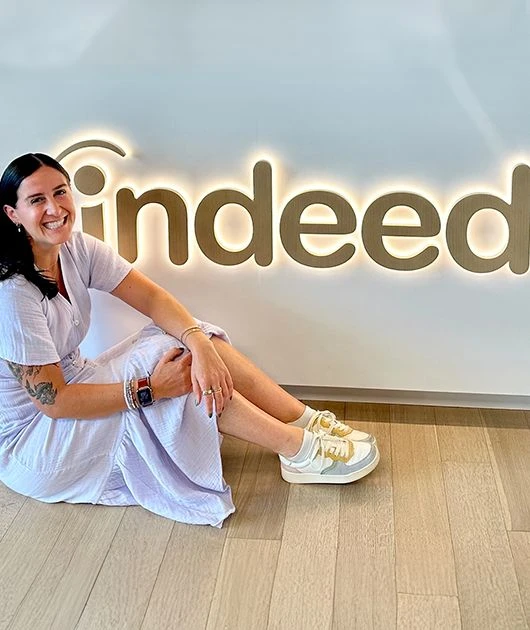 Person sitting on the floor beside a lit Indeed sign on the wall, smiling at the camera