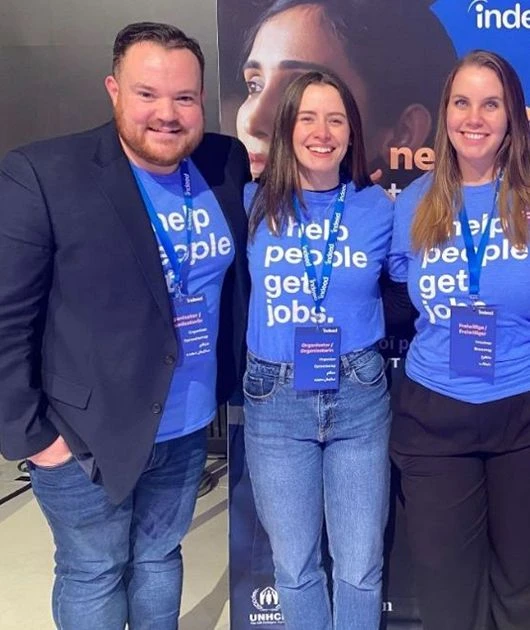 Three individuals wearing blue t-shirts with Indeed logos and the phrase We help people get jobs stand together at an event.