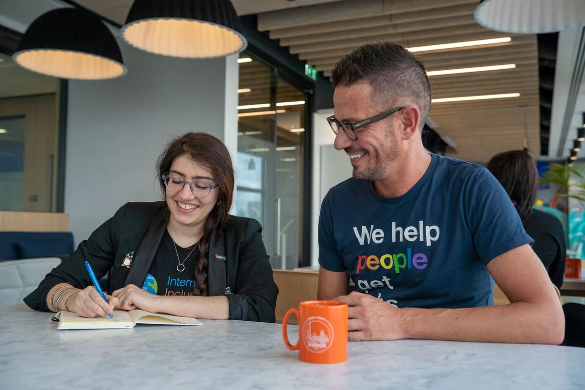 Two people in a modern office setting smiling and discussing over a notebook with an orange mug on the table.