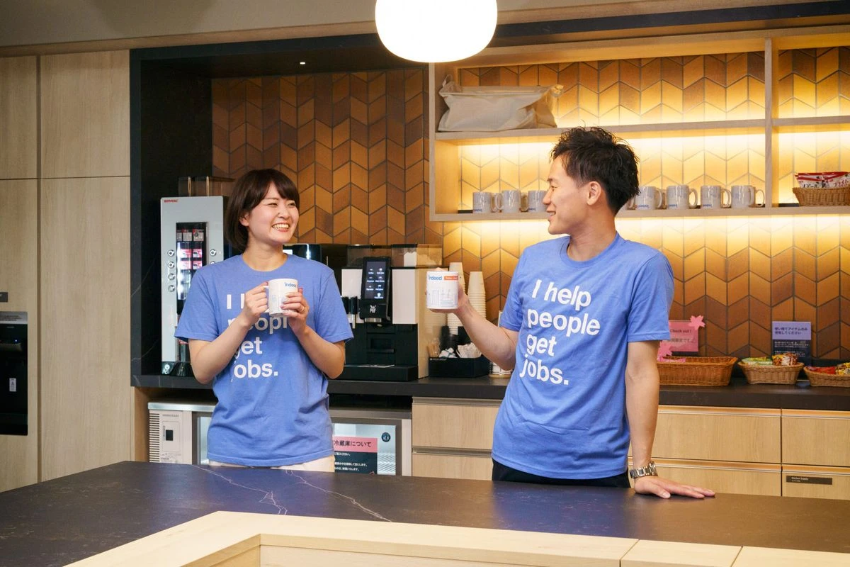 Two individuals wearing blue t-shirts with the text I help people get jobs. stand in a modern kitchen, smiling and holding mugs.