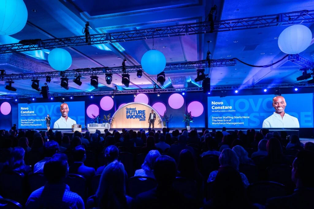 From the back of the conference room, you can see hte entire audience watching Indeed GM of Stafing Solutions and Indeed Flex Novo Constare standing in the middle of the FutureWorks stage. He is a black man with a shaved head in a dark suit, and beside him is his introduction slide of his presentation featuring his headshot.