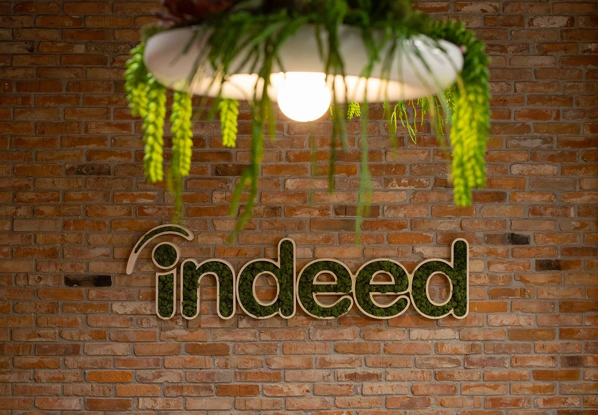 A lit hanging lamp with green plants in front of a brick wall with the Indeed logo