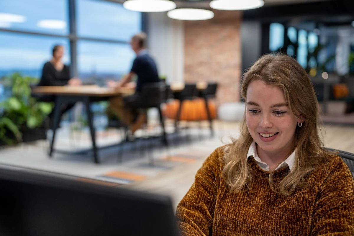 A person smiling while working on a computer in a modern office, with two colleagues visible in the background engaged in discussion