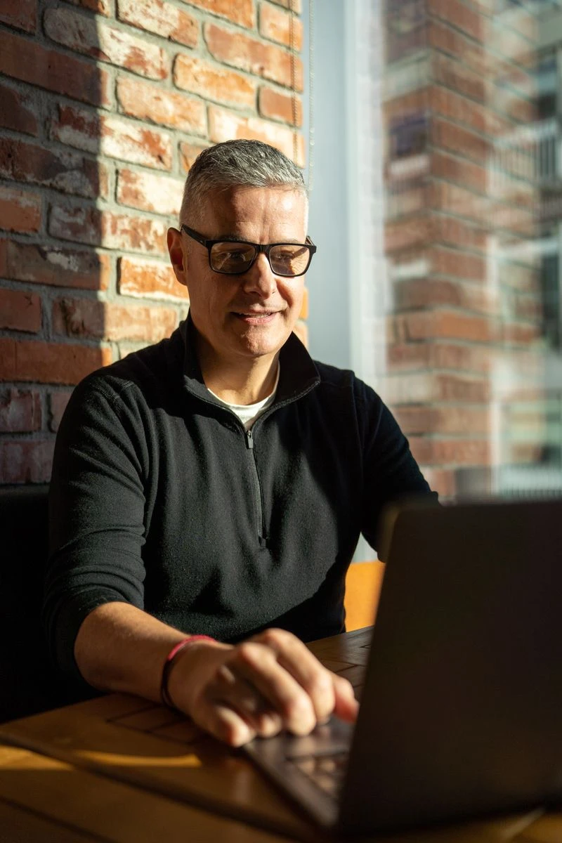 A man with short grey hair and glasses sits at a table in front of a brick wall, illuminated by sunlight. He is wearing a black collared shirt with a zipper and is looking down at a laptop in front of him.