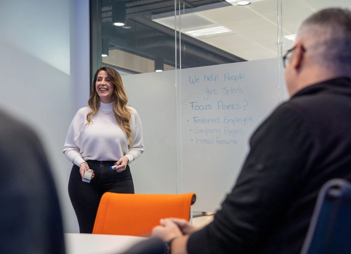 A smiling woman with long, wavy brown hair stands in front of a whiteboard, holding a blue marker. She is wearing a white ribbed sweater and black pants. In the foreground, the back of a man's head and shoulder are visible, along with an orange office chair.