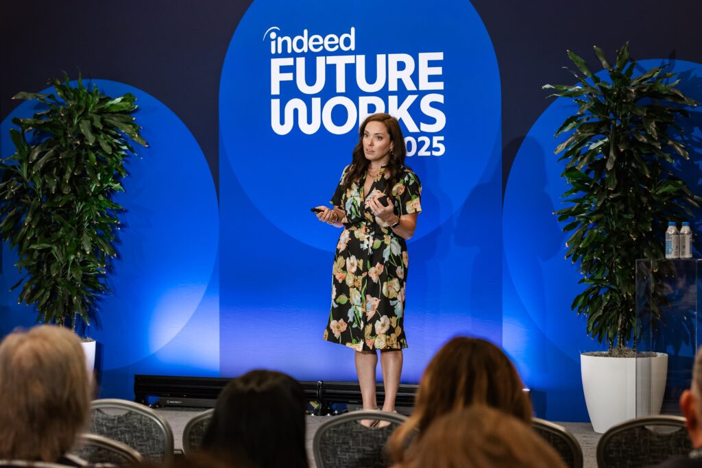 Indeed Global Client Strategist Linsey Fagan speaks on stage at FutureWorks 2025 in front of an audience of employers. She is a white woman with long dark hair, wearing hoop earrings and a black dress with a floral print.