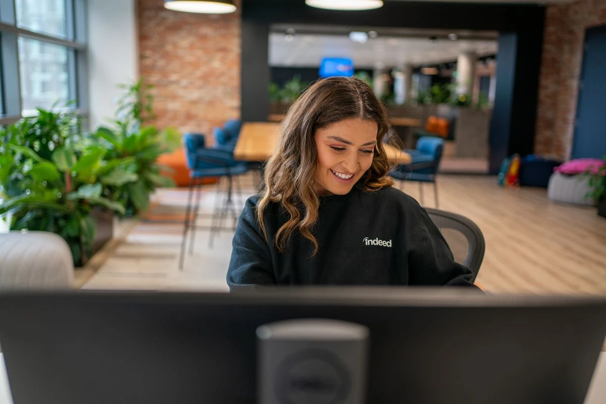 A smiling person wearing a black long-sleeved shirt with Indeed logo written in white text on the front. In the blurred background, an office space.